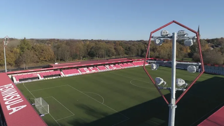 Đèn pha chiếu sáng rực rỡ tại sân vận động Peninsula Stadium vào buổi tối, tạo nên không khí trận đấu bóng đá